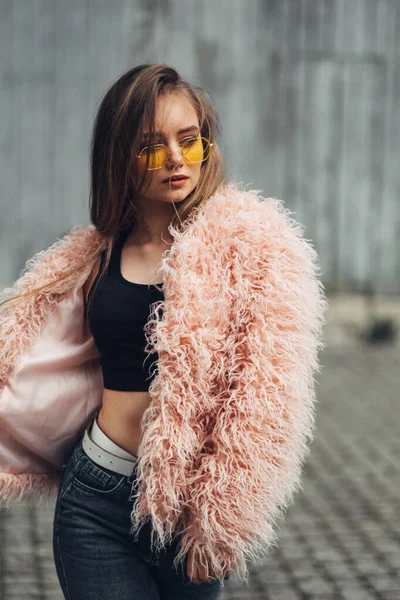 Portret van een stijlvolle meisje in roze vacht en moderne gele zonnebril poseren op straat — Stockfoto