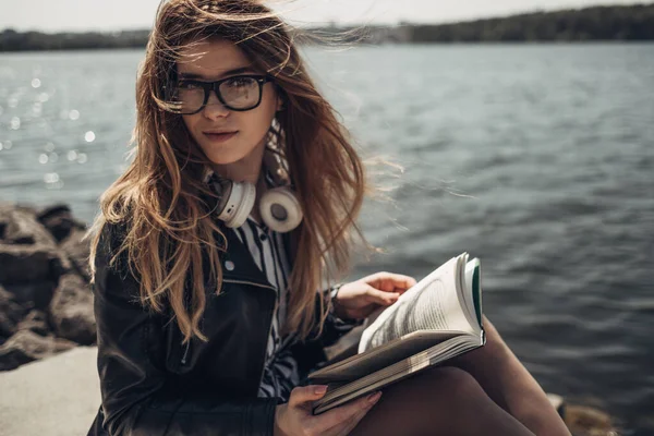 Retrato de verão ao ar livre da menina bonita nova em jaqueta de couro preto e óculos lê o livro perto do lago — Fotografia de Stock