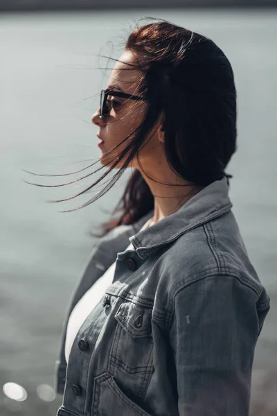 Schöne brünette Mädchen mit Sonnenbrille weiße Hose und Jeansjacke in der Nähe des Sees — Stockfoto