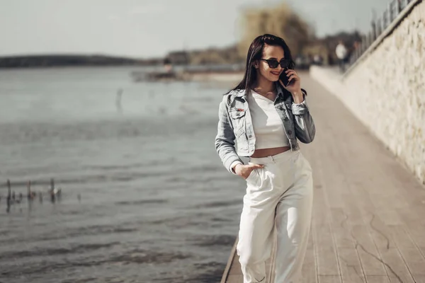 Piękna brunetka dziewczyna w okulary przeciwsłoneczne białe spodnie i dżinsy kurtka w pobliżu jeziora — Zdjęcie stockowe