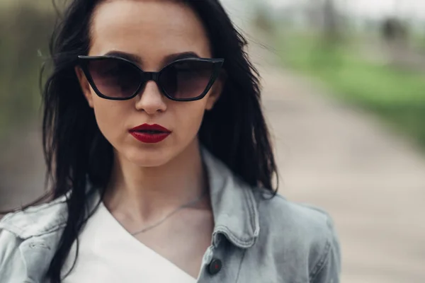 Schöne brünette Mädchen mit Sonnenbrille, weißer Hose und Jeansjacke, rotem Lippenstift — Stockfoto
