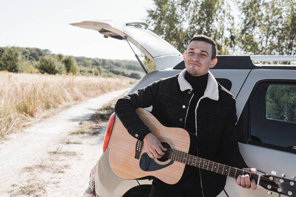 Portrait of One Man Dressed in Black Stylish Clothes Enjoying a Car Trip on the Field Road, Playing Guitar, Vacation Concept