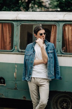 Güneş gözlüklü, kot ceketli ve eski arabanın yanında beyaz tişörtlü şık bir çocuğun yakın portresi.