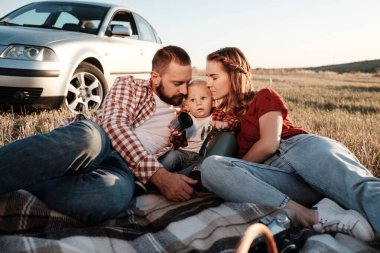 Mutlu Genç Aile Anne ve Baba ve Küçük Oğulları Hafta Sonu Keyifli Pikniği. Ekoseli Ekoseli Arabanın Üzerinde, Şehrin Dışında, Sunny Day Sunset, Tatil ve Yolculuk Konsepti 'nde.