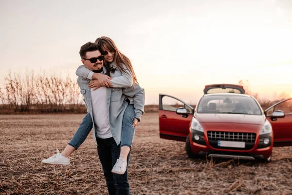 Jovem casal feliz vestido de igual em camisa branca e jeans desfrutando de viagem rodoviária em seu novo carro, belo pôr do sol no campo, férias e conceito de viagem — Fotografia de Stock