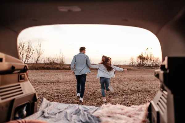 Jovem casal feliz vestido igual em camisa branca e jeans sentado em seu novo tronco de carro, belo pôr do sol no campo, férias e conceito de viagem — Fotografia de Stock