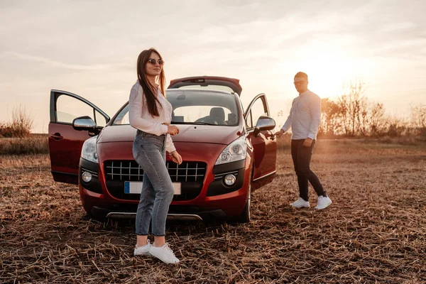 Young Happy Couple Dressed Alike in White Shirt and Jeans Enjoying Road Trip at Their New Car, Beautiful Sunset on the Field, Vacation and Travel Concept — Stock Photo, Image