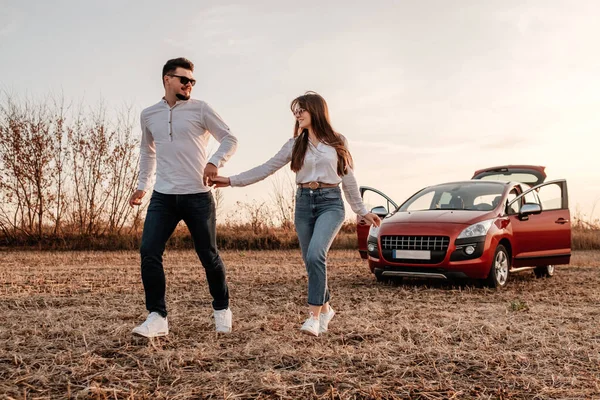 Joven pareja feliz vestida como en camisa blanca y pantalones vaqueros disfrutando de un viaje por carretera en su nuevo coche, hermoso atardecer en el campo, vacaciones y concepto de viaje —  Fotos de Stock