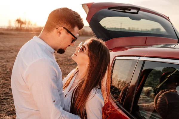 Joven pareja feliz vestida como en camisa blanca y pantalones vaqueros disfrutando de un viaje por carretera en su nuevo coche, hermoso atardecer en el campo, vacaciones y concepto de viaje — Foto de Stock