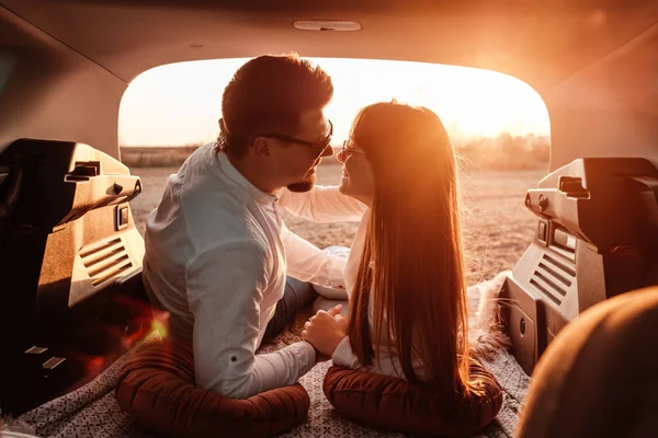 Jovem casal feliz vestido de igual em camisa branca e jeans desfrutando de viagem rodoviária em seu novo carro, belo pôr do sol no campo, férias e conceito de viagem — Fotografia de Stock