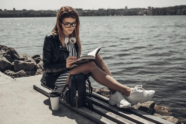 Retrato de verão ao ar livre da menina bonita nova em jaqueta de couro preto e óculos lê o livro perto do lago — Fotografia de Stock