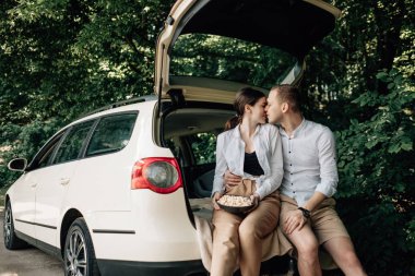 Beyaz tişörtlü genç mutlu çift yol kenarında patlamış mısırlı arabada oturuyor, haftasonu şehrin dışında, tatiller ve yolculuk konsepti.