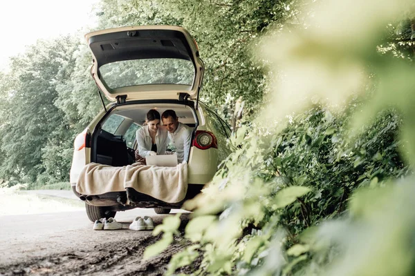 Joven Pareja Feliz Vestida Como en Camiseta Blanca Sentada en el Maletero del Coche con Portátil y Palomitas de Maíz en la Carretera, Fin de Semana Fuera de la Ciudad, Vacaciones y Concepto de Viaje por Carretera — Foto de Stock