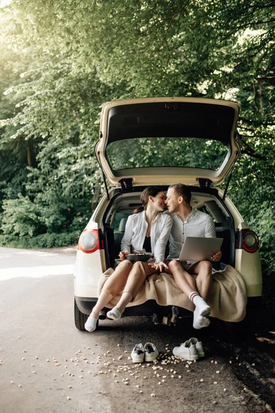 Joven Pareja Feliz Vestida Como en Camiseta Blanca Sentada en el Maletero del Coche con Portátil y Palomitas de Maíz en la Carretera, Fin de Semana Fuera de la Ciudad, Vacaciones y Concepto de Viaje por Carretera — Foto de Stock