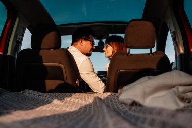 Beyaz gömlekli ve kot pantolonlu genç mutlu çift yeni arabalarında yolculuğun tadını çıkarıyor, Sahada güzel bir gün batımı, tatil ve seyahat konsepti