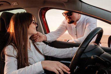 Beyaz gömlekli ve kot pantolonlu genç mutlu çift yeni arabalarında yolculuğun tadını çıkarıyor, Sahada güzel bir gün batımı, tatil ve seyahat konsepti