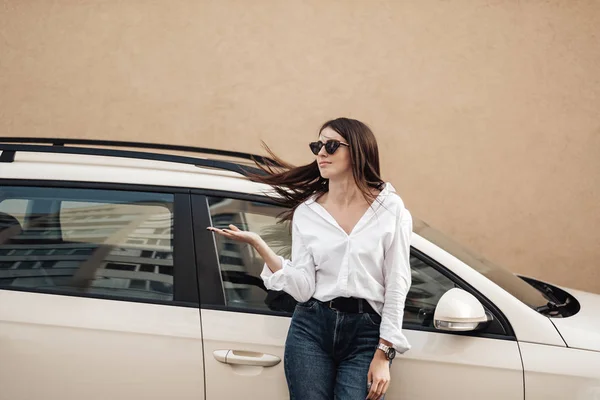 Primer plano Retrato de una joven elegante vestida con pantalones vaqueros y camisa blanca posando cerca del coche, dama de negocios, concepto de poder de la mujer — Foto de Stock