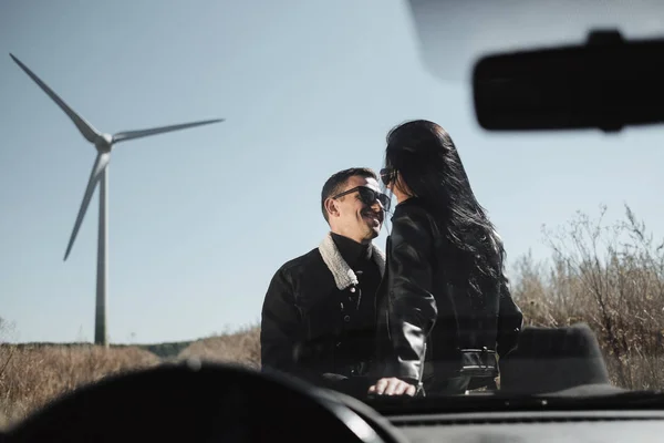 Happy Traveling Couple Enjoying a Car Trip on the Field Road with Electric Wind Turbine Power Generator on the Background