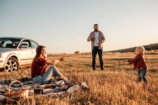 Heureux jeune famille maman et papa avec leur petit fils profiter week-end d'été pique-nique sur la voiture en dehors de la ville, jouer avec des bulles — Photo