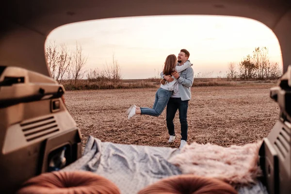 Jovem casal feliz vestido igual em camisa branca e jeans sentado em seu novo tronco de carro, belo pôr do sol no campo, férias e conceito de viagem — Fotografia de Stock