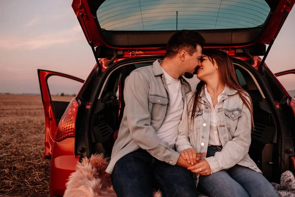 Young Happy Couple Dressed Alike in White Shirt and Jeans Sitting at Their New Car Trunk, Beautiful Sunset on the Field, Vacation and Travel Concept — Stock Photo, Image