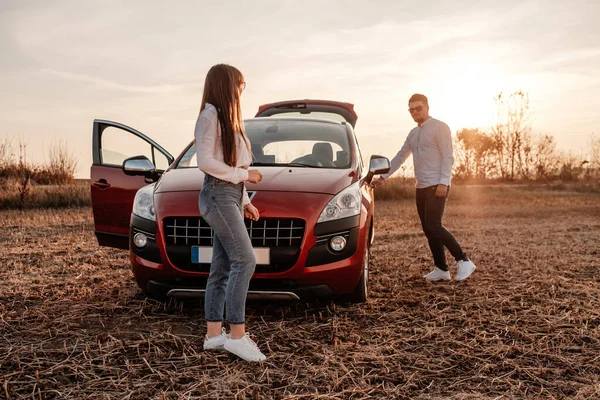 Young Happy Couple Dressed Alike in White Shirt and Jeans Enjoying Road Trip at Their New Car, Beautiful Sunset on the Field, Vacation and Travel Concept — Stock Photo, Image