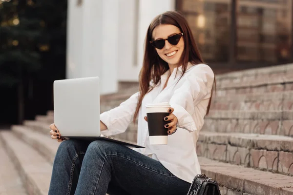 Retrato de uma menina elegante vestida em jeans e camisa branca segurando laptop e café quente, trabalhador freelance, senhora de negócios, conceito de poder da mulher — Fotografia de Stock