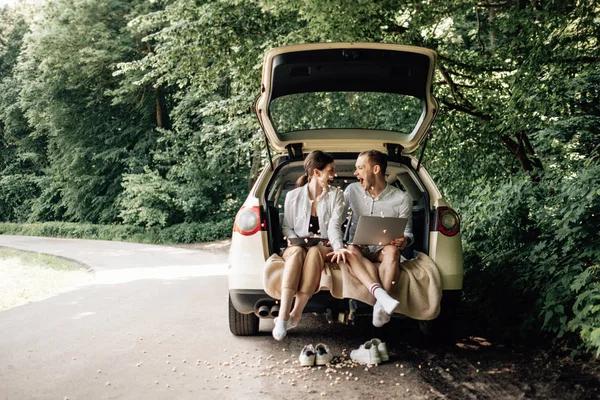 Young Happy Couple Dressed Alike in White T-shirt Sitting in the Car Trunk with Laptop and Popcorn on the Roadside, Weekend Outside the City, Holidays and Road Trip Concept — стокове фото