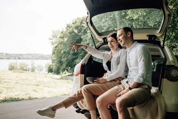 Jeune couple heureux habillé comme dans un t-shirt blanc assis dans le coffre de la voiture avec du maïs soufflé sur la route, week-end hors de la ville, vacances et voyage concept — Photo