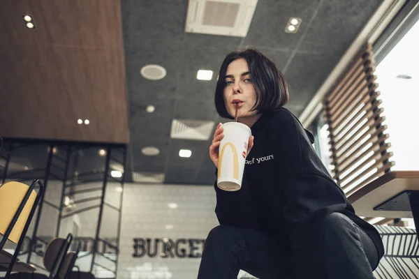 TERNOPIL, UCRAINA - 25 MAGGIO 2019: Bella ragazza bruna in Pullover nero con testo "Best Version of Yourself" beve Coca-Cola in McDonalds — Foto Stock