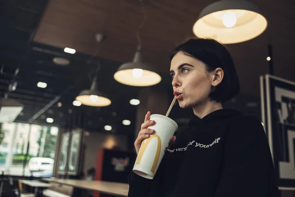 TERNOPIL, UCRAINA - 25 MAGGIO 2019: Bella ragazza bruna in Pullover nero con testo "Best Version of Yourself" beve Coca-Cola in McDonalds — Foto Stock