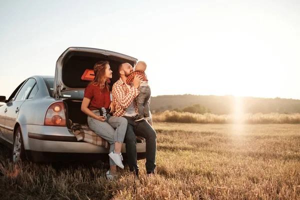 Glad ung familj mamma och pappa med sin lilla son Njuter av sommaren Weekend picknick Sitter på bagageluckan av bilen utanför staden i fältet på soliga dagen solnedgång, semester och väg resa koncept — Stockfoto