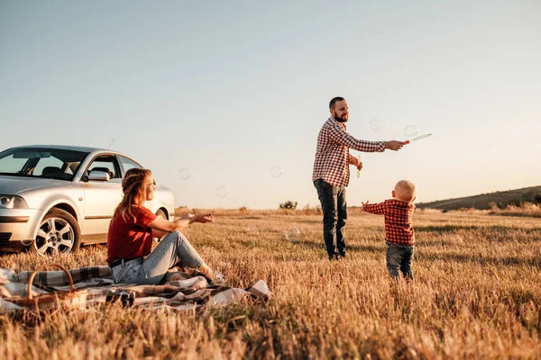 Gelukkig jong gezin mam en pap met hun kleine zoon genieten van de zomer weekend picknick op de auto buiten de stad, spelen met bubbels — Stockfoto
