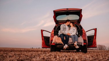 Beyaz Gömlekli ve Kot Giyen Genç Mutlu Çift Yeni Araba Bagajlarında Oturuyor, Sahada Güzel Günbatımı, Tatil ve Seyahat Konsepti