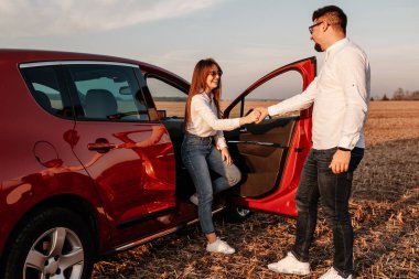 Beyaz gömlekli ve kot pantolonlu genç mutlu çift yeni arabalarında yolculuğun tadını çıkarıyor, Sahada güzel bir gün batımı, tatil ve seyahat konsepti