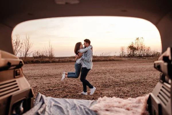 Jovem casal feliz vestido igual em camisa branca e jeans sentado em seu novo tronco de carro, belo pôr do sol no campo, férias e conceito de viagem — Fotografia de Stock