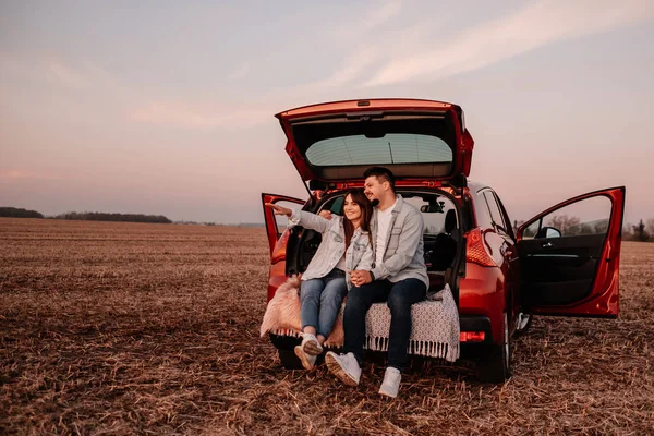 Joven pareja feliz vestida como en camisa blanca y pantalones vaqueros sentados en su nuevo baúl de coche, hermoso atardecer en el campo, vacaciones y concepto de viaje —  Fotos de Stock