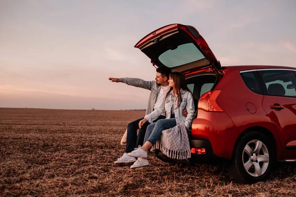 Young Happy Couple Dressed Alike in White Shirt and Jeans Sitting at Their New Car Trunk, Beautiful Sunset on the Field, Vacation and Travel Concept — 스톡 사진