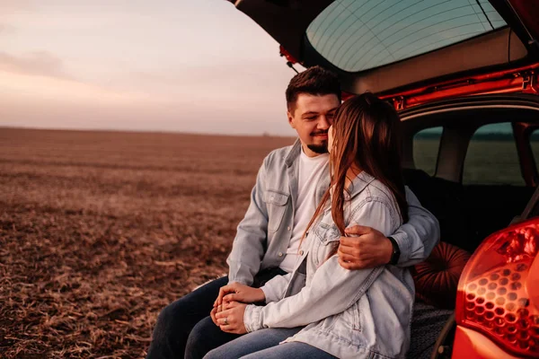 Jovem casal feliz vestido igual em camisa branca e jeans sentado em seu novo tronco de carro, belo pôr do sol no campo, férias e conceito de viagem — Fotografia de Stock