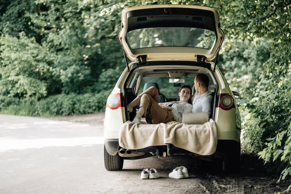 Joven Pareja Feliz Vestida Como en Camiseta Blanca Sentada en el Maletero del Coche con Portátil y Palomitas de Maíz en la Carretera, Fin de Semana Fuera de la Ciudad, Vacaciones y Concepto de Viaje por Carretera — Foto de Stock