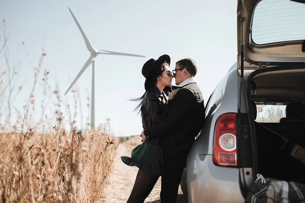 Счастливая путешествующая пара наслаждается поездкой на автомобиле по полевой дороге с электрическим генератором ветровой турбины на заднем плане — стоковое фото