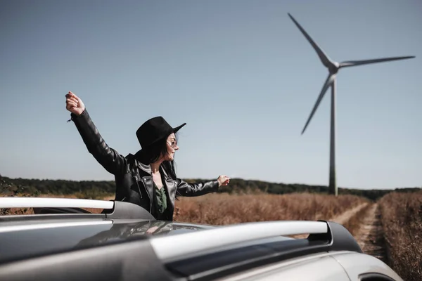 Happy Traveling Girl Enjoying a Car Trip on the Field Road with Electric Wind Turbine Power Generator on the Background