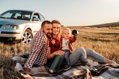 Mutlu Genç Aile Anne ve Baba ve Küçük Oğulları Hafta Sonu Keyifli Pikniği. Ekoseli Ekoseli Arabanın Üzerinde, Şehrin Dışında, Sunny Day Sunset, Tatil ve Yolculuk Konsepti 'nde.