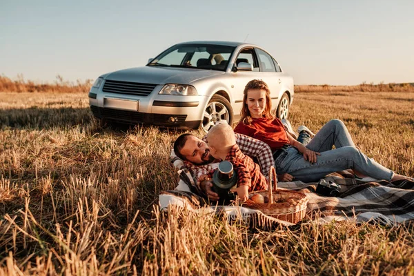 小さな息子と幸せな若い家族のお母さんとお父さん夏の週末のピクニックを楽しむ晴れた日の日没、休暇、ロードトリップのコンセプトで市外の車の近くに座って — ストック写真