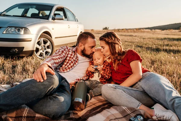 Happy Young Family Maman et papa avec leur petit fils profiter week-end d'été pique-nique assis sur la plaid près de la voiture à l'extérieur de la ville dans le champ au coucher du soleil, vacances et voyage concept — Photo