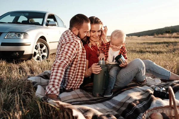 Glad ung familj mamma och pappa med sin lilla son Njuter av sommaren Weekend picknick Sitter på rutig nära bilen utanför staden på fältet på soliga dagen solnedgång, semester och väg resa koncept — Stockfoto