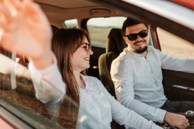 Beyaz gömlekli ve kot pantolonlu genç mutlu çift yeni arabalarında yolculuğun tadını çıkarıyor, Sahada güzel bir gün batımı, tatil ve seyahat konsepti