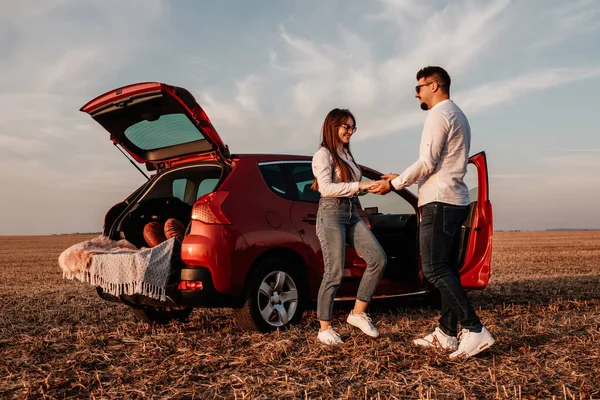 Joven pareja feliz vestida como en camisa blanca y pantalones vaqueros disfrutando de un viaje por carretera en su nuevo coche, hermoso atardecer en el campo, vacaciones y concepto de viaje —  Fotos de Stock