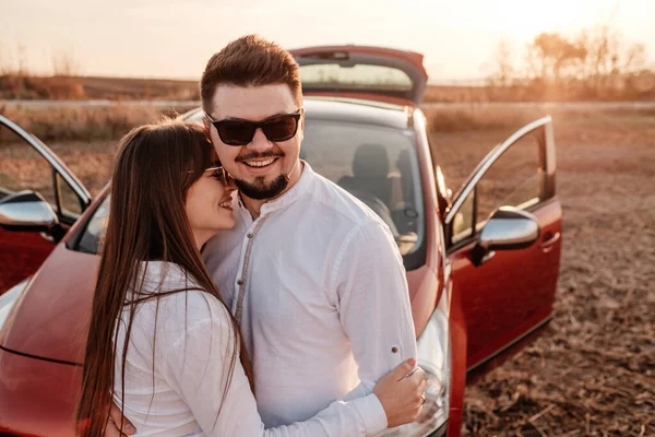 Jovem casal feliz vestido de igual em camisa branca e jeans desfrutando de viagem rodoviária em seu novo carro, belo pôr do sol no campo, férias e conceito de viagem — Fotografia de Stock