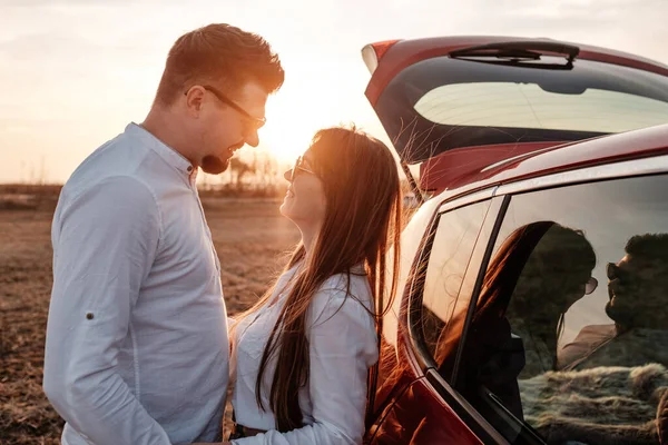 Joven pareja feliz vestida como en camisa blanca y pantalones vaqueros disfrutando de un viaje por carretera en su nuevo coche, hermoso atardecer en el campo, vacaciones y concepto de viaje —  Fotos de Stock